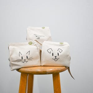 May include: Three beige canvas bags with black line drawings of a cat, dog, and dinosaur. The bags are on a wooden stool against a white wall. Each bag has a small green circular tag.