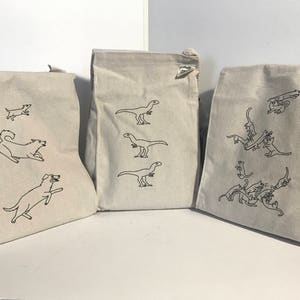 May include: Three white canvas tote bags with black line drawings of dogs and dinosaurs. The bag on the left has a dog running and jumping. The middle bag has three dinosaurs in different poses. The bag on the right has a group of cats playing.