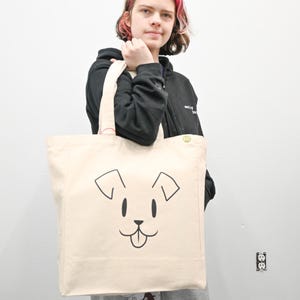 May include: A person is holding a beige canvas tote bag with a black and white cartoon dog illustration. The dog has a big smile and its tongue is sticking out.