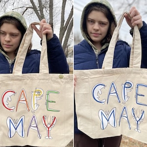 May include: A beige tote bag with the words "CAPE MAY" in colorful, whimsical lettering. The letters are designed with marine life illustrations, including a shark, crab, butterfly, and lobster. The bag has sturdy handles.