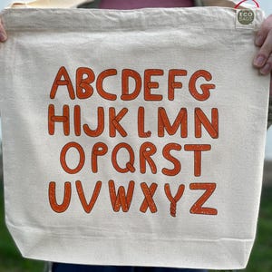 May include: A beige canvas tote bag with the alphabet printed in orange letters. The letters are in a bold, sans-serif font. The bag has a simple design and is held by a person.