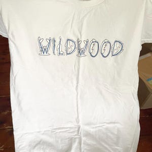 May include: White t-shirt with the word "WILDWOOD" in blue, stylized with whimsical illustrations. The shirt is made of a soft, likely cotton material, and has a classic crew neck design. The text is centered on the front of the shirt.