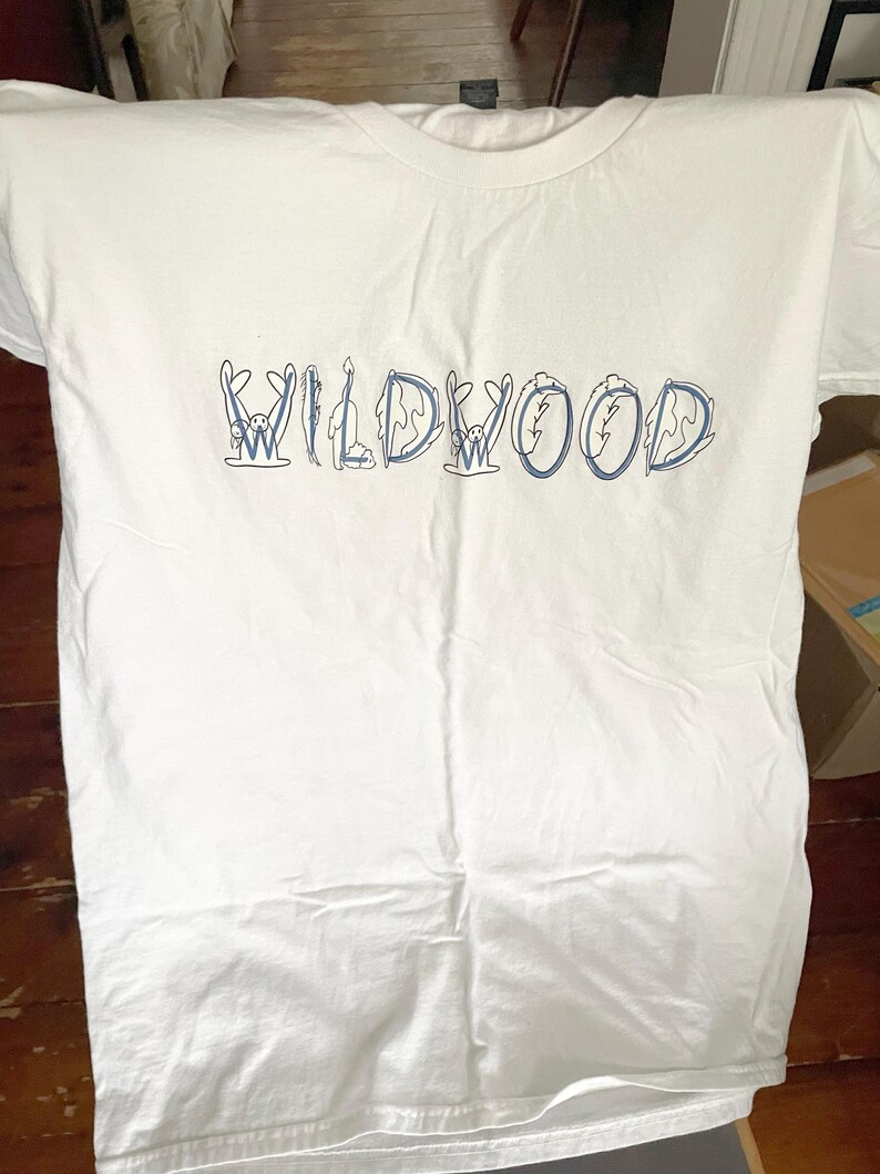 May include: White t-shirt with the word "WILDWOOD" in blue, stylized with whimsical illustrations. The shirt is made of a soft, likely cotton material, and has a classic crew neck design. The text is centered on the front of the shirt.