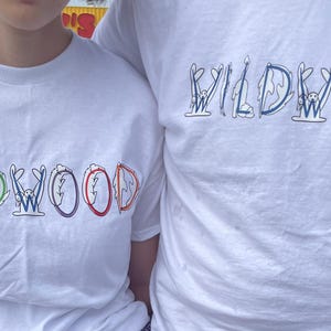 May include: White t-shirts with the word "WILDWOOD" in colorful, whimsical lettering. The letters are designed with various illustrations, including a bunny, a flame, and a wave. The shirts are short-sleeved.