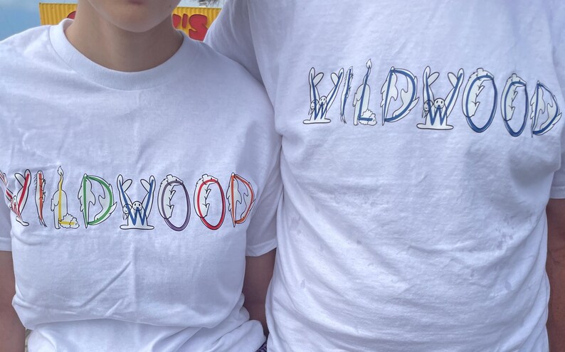 May include: White t-shirts with the word "WILDWOOD" in colorful, whimsical lettering. The letters are designed with various illustrations, including a bunny, a flame, and a wave. The shirts are short-sleeved.