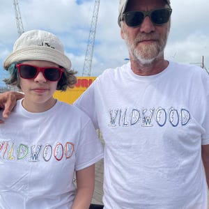 May include: Two people wearing white t-shirts with the word "WILDWOOD" in colorful, stylized lettering. The person on the left is wearing a white bucket hat and red sunglasses. The person on the right is wearing a baseball cap and sunglasses.