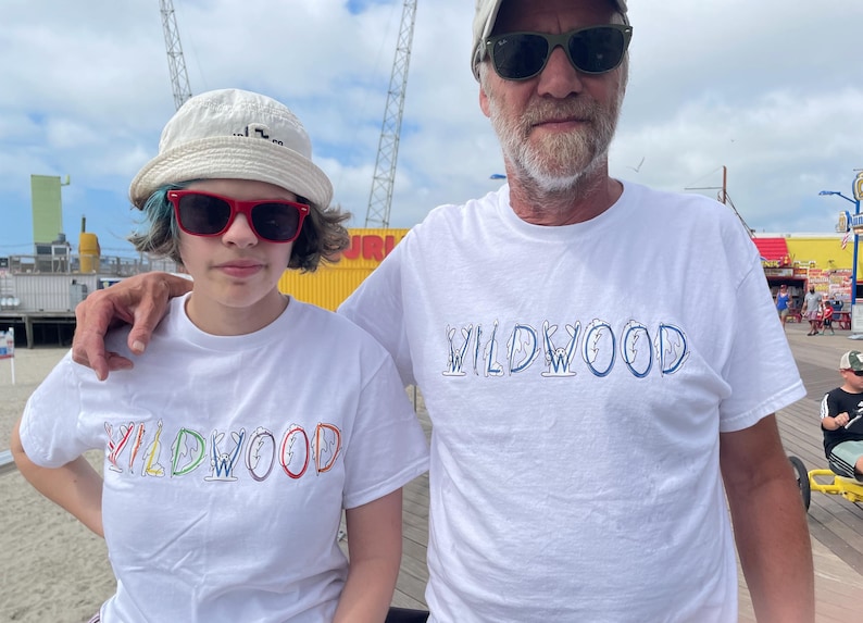 May include: Two people wearing white t-shirts with the word "WILDWOOD" in colorful, stylized lettering. The person on the left is wearing a white bucket hat and red sunglasses. The person on the right is wearing a baseball cap and sunglasses.