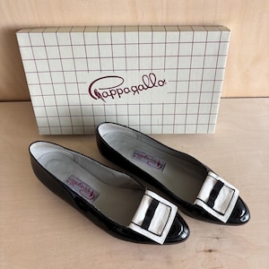 Könnte beinhalten: Ein Paar flache Schuhe aus schwarzem Lackleder mit einer weißen rechteckigen Schnalle und einem schwarzen Mitteldetail. Die Schuhe stehen vor einem weißen Karton mit braunem Gittermuster und dem Markennamen Pappagallo in brauner Schreibschrift.