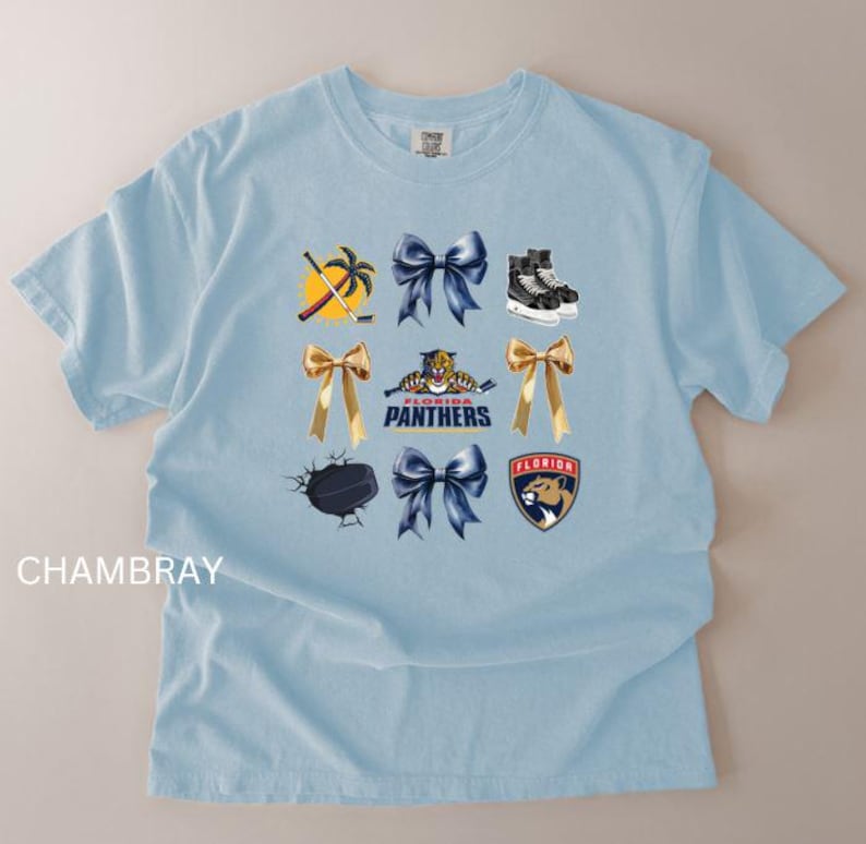 May include: Light blue t-shirt with a variety of graphics, including hockey-themed images, bows, and the words "Florida Panthers". The shirt has a crew neck and short sleeves. The word "CHAMBRAY" is printed on the left side of the shirt.