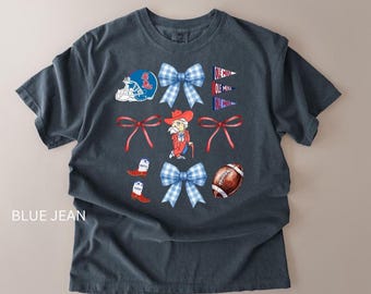 May include: Blue jean colored t-shirt with a graphic design featuring a football helmet, bows, a cartoon character, football, and boots. The design incorporates red, white, and blue colors, with the text "OLE MISS".