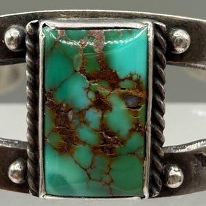 May include: A silver cuff bracelet featuring a rectangular turquoise stone with brown matrix in the center. The metal band is decorated with stamped chevron patterns and small silver spheres on either side of the stone setting.