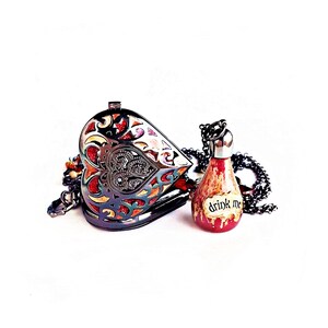 May include: A heart-shaped, ornate metal pendant with colorful stained glass-like accents. Next to it is a small, red potion bottle necklace with the words "drink me" on a label. Both are on a white background.