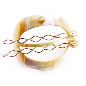 May include: A set of three gold hair clips with a wavy design. The clips are attached to a white, pearlescent, oval-shaped base.
