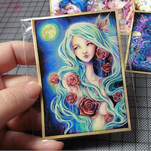 May include: A woman with long, flowing blue-green hair is surrounded by red roses. The background is a dark blue with a full moon and stars. The image has a dreamy, ethereal feel.