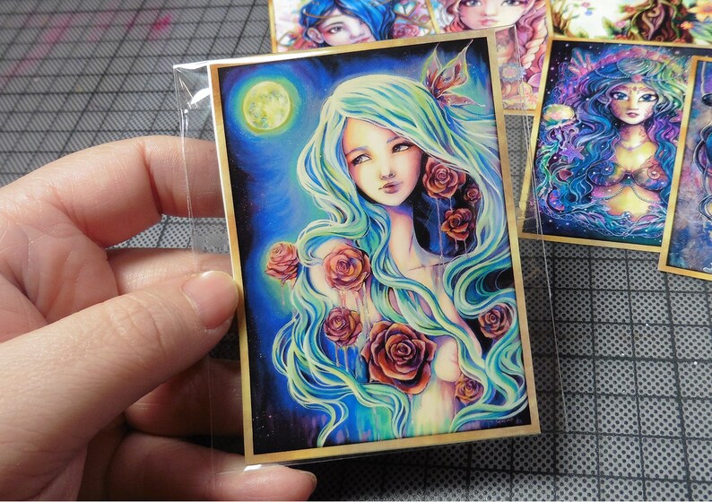 May include: A woman with long, flowing blue-green hair is surrounded by red roses. The background is a dark blue with a full moon and stars. The image has a dreamy, ethereal feel.