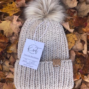 May include: A beige knit beanie with a faux fur pom-pom. The hat has a folded brim and a tag that reads "Fold Over Brim Beanie" and "Hooks & Heart". The hat is displayed on a bed of autumn leaves.