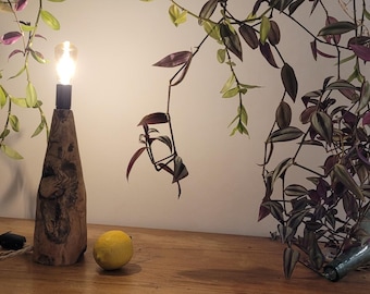 May include: A wooden lamp with a light bulb on top, sitting on a wooden table with two lemons in front of it. A plant with purple and green leaves hangs over the table.