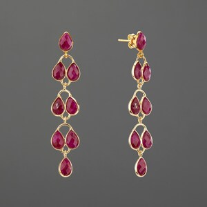 May include: A pair of gold drop earrings featuring multiple faceted, teardrop-shaped magenta gemstones arranged in a cascading vertical design. Each earring is suspended from a post back and consists of several linked stones in a gold setting.