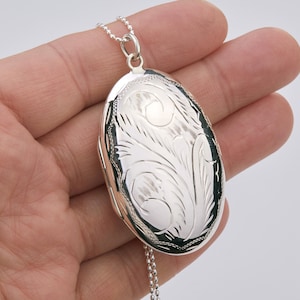 May include: Oval silver locket pendant with detailed floral engraving, suspended from a fine silver chain. The locket is about 3.8 cm long and has a hinged design for holding small keepsakes.