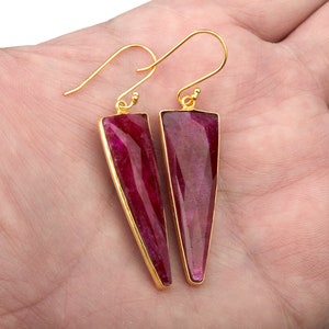 Op de afbeelding: Een paar goudkleurige oorbellen met driehoekige, dieprode edelstenen. De edelstenen zijn gezet in een gouden montuur en hebben een gepolijste, facetgeslepen uitstraling. De oorbellen hebben een haaksluiting.