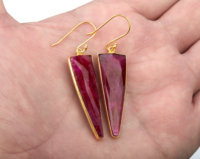 Op de afbeelding: Een paar goudkleurige oorbellen met driehoekige, dieprode edelstenen. De edelstenen zijn gezet in een gouden montuur en hebben een gepolijste, facetgeslepen uitstraling. De oorbellen hebben een haaksluiting.