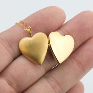May include: A gold-colored heart-shaped locket pendant on a delicate gold chain. The locket is open, revealing a matte, brushed gold finish on the exterior left side and a smooth, highly polished, reflective gold finish on the interior right side.