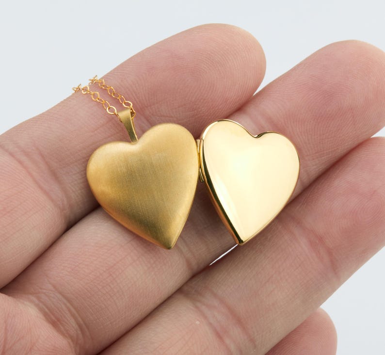 May include: A gold-colored heart-shaped locket pendant on a delicate gold chain. The locket is open, revealing a matte, brushed gold finish on the exterior left side and a smooth, highly polished, reflective gold finish on the interior right side.