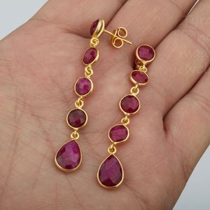 May include: A pair of gold earrings with dangling red gemstone drops. Each earring has five round red gemstones and a teardrop-shaped gemstone at the bottom.
