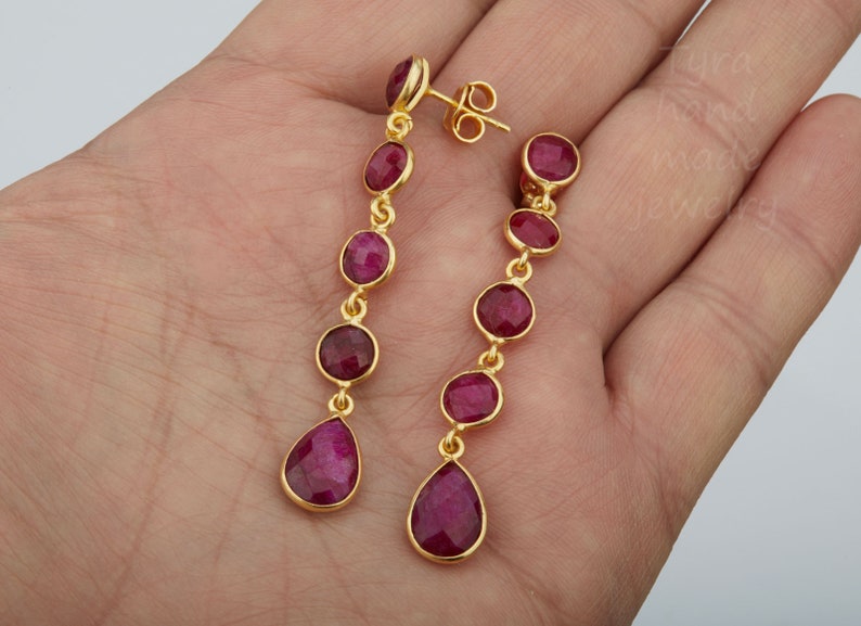 May include: A pair of gold earrings with dangling red gemstone drops. Each earring has five round red gemstones and a teardrop-shaped gemstone at the bottom.