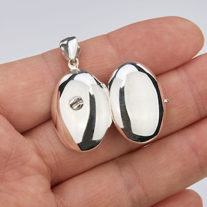 Könnte beinhalten: Ein silberner ovaler Anhänger mit einer Scharnieröffnung. Das Medaillon ist geöffnet und zeigt ein glattes, leeres Inneres.