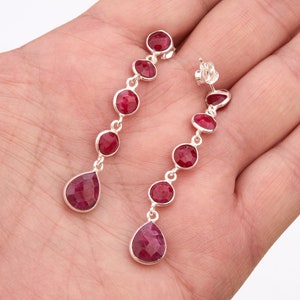 May include: A pair of silver dangle earrings with red gemstone drops. The earrings feature a row of round red gemstones and a teardrop-shaped red gemstone at the bottom.