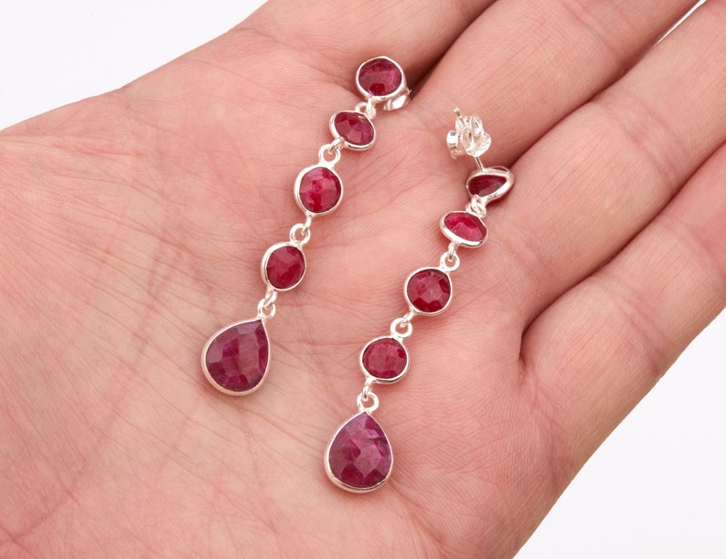 May include: A pair of silver dangle earrings with red gemstone drops. The earrings feature a row of round red gemstones and a teardrop-shaped red gemstone at the bottom.
