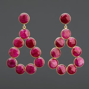 Puede incluir: Un par de pendientes colgantes con forma de lágrima. Los pendientes están hechos de oro y presentan un grupo de piedras preciosas redondas de color rosa oscuro.