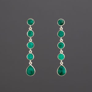 May include: A pair of silver dangle earrings with a teardrop-shaped emerald gemstone at the bottom and five round emerald gemstones above it.