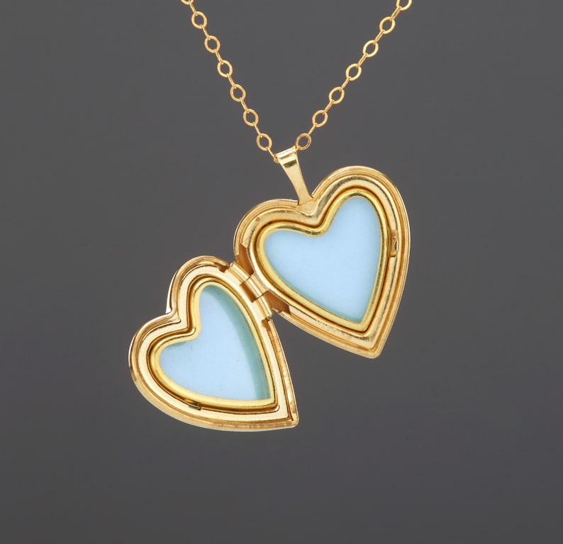 May include: A gold-toned heart-shaped locket pendant suspended from a matching delicate chain. The locket is open, revealing two empty heart-shaped compartments with smooth, light blue interior surfaces framed by polished gold-colored metal edges.