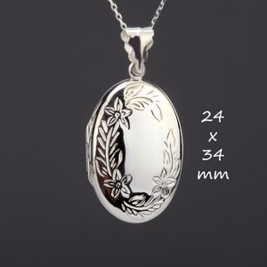 Puede incluir: Colgante de plata con forma de relicario ovalado con detalles florales, suspendido de una delicada cadena. El relicario mide 24 x 34 mm. El diseño presenta flores y hojas grabadas en los bordes. El fondo es gris oscuro.