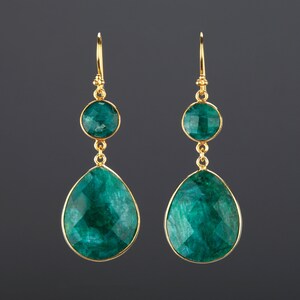 May include: A pair of gold earrings with two green gemstone drops. The top gemstone is round and the bottom gemstone is teardrop shaped.