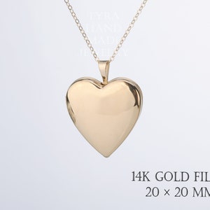 May include: A gold-colored heart-shaped locket pendant on a delicate chain. The locket is marked 14K GOLD FILLED 20 x 20 MM. The text TYRA HAND MADE JEWELLERY is visible in the background. The surface of the heart is smooth and reflective.