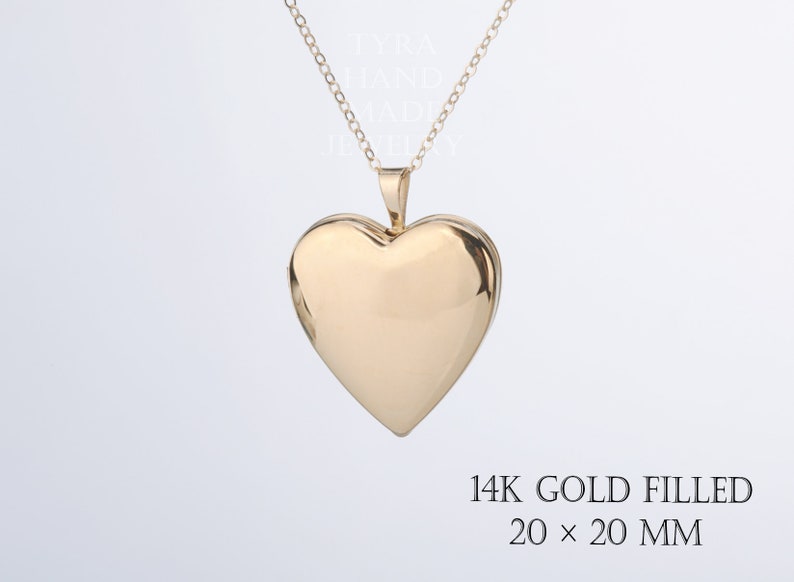 May include: A gold-colored heart-shaped locket pendant on a delicate chain. The locket is marked 14K GOLD FILLED 20 x 20 MM. The text TYRA HAND MADE JEWELLERY is visible in the background. The surface of the heart is smooth and reflective.