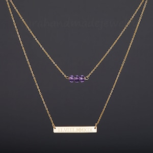 Puede incluir: Un collar de cadena dorada con un colgante de barra grabado con los números romanos "II.VIII.MMXIX" y una cadena más pequeña con una fila de cuentas moradas.