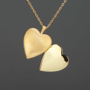 May include: A gold heart-shaped locket pendant suspended from a delicate gold chain. The locket is open, revealing a matte finish on the left side and a polished, reflective surface on the right side, showcasing a classic hinged design.
