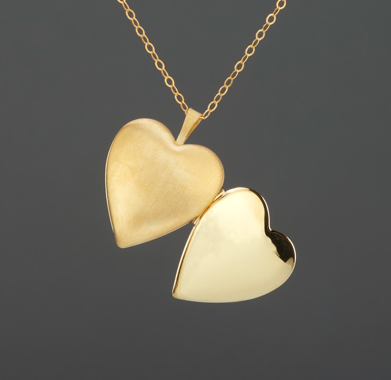 May include: A gold heart-shaped locket pendant suspended from a delicate gold chain. The locket is open, revealing a matte finish on the left side and a polished, reflective surface on the right side, showcasing a classic hinged design.