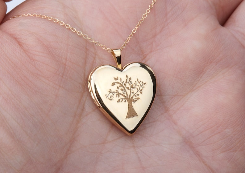 May include: A gold-toned heart-shaped locket pendant suspended from a delicate matching chain. The front surface of the locket features a detailed, dark-etched engraving of a tree with spreading branches and leaves centered on the polished metal.