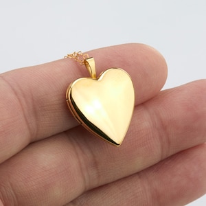 May include: A polished gold-tone heart-shaped locket pendant suspended from a delicate matching gold-tone chain. The locket features a smooth, reflective surface with a classic heart silhouette and a small bail at the top for the chain attachment.