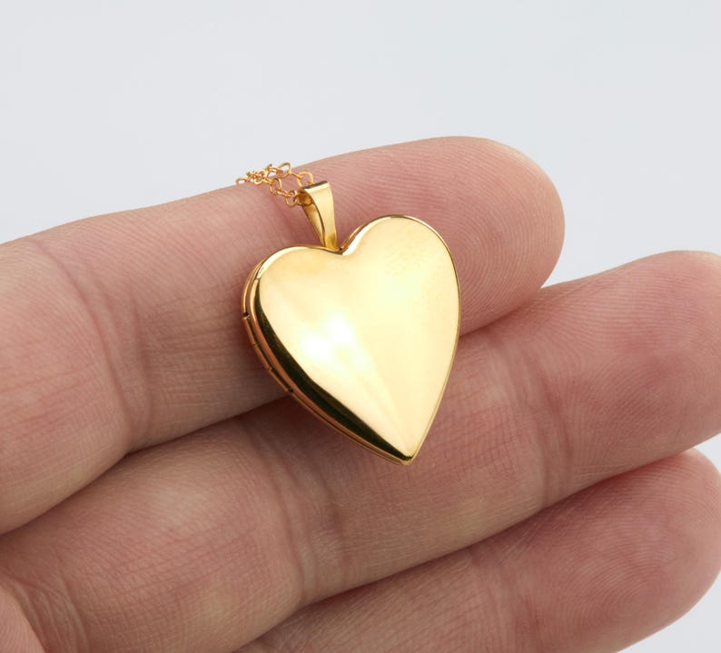 May include: A polished gold-tone heart-shaped locket pendant suspended from a delicate matching gold-tone chain. The locket features a smooth, reflective surface with a classic heart silhouette and a small bail at the top for the chain attachment.