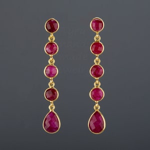 May include: A pair of gold earrings with a teardrop-shaped ruby gemstone at the bottom and four round ruby gemstones above it.