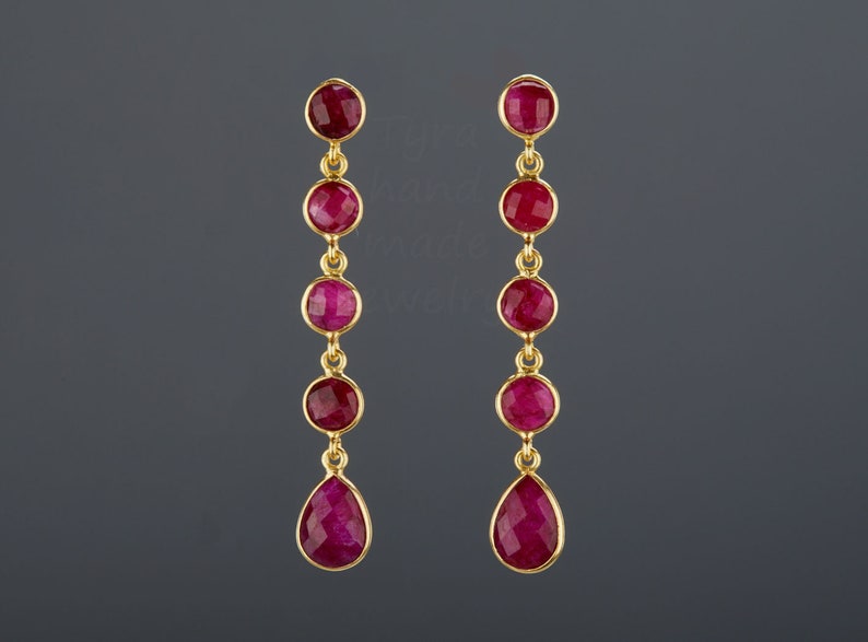 May include: A pair of gold earrings with a teardrop-shaped ruby gemstone at the bottom and four round ruby gemstones above it.