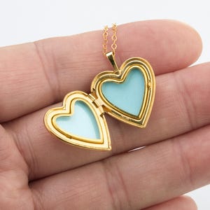 May include: A gold-toned heart-shaped locket pendant on a delicate matching chain. The locket is open, revealing two empty, light blue interior compartments framed by polished gold-colored metal edges. The piece features a classic hinged design.