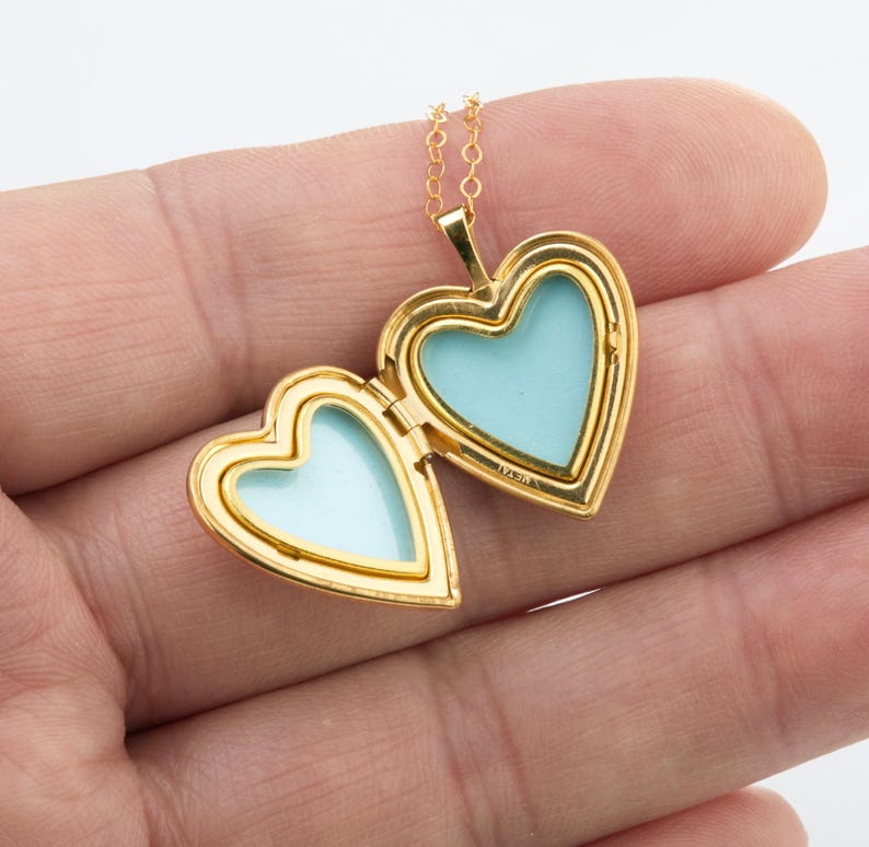 May include: A gold-toned heart-shaped locket pendant on a delicate matching chain. The locket is open, revealing two empty, light blue interior compartments framed by polished gold-colored metal edges. The piece features a classic hinged design.