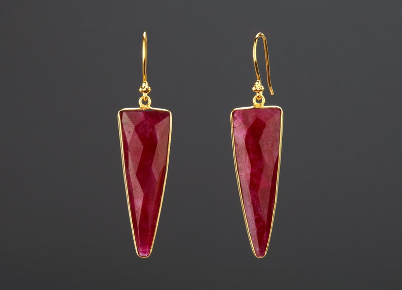 Op de afbeelding: Een paar goudkleurige oorhangers met grote, facetgeslepen, driehoekige robijnrode edelstenen. De oorbellen hebben een eenvoudig ontwerp met een haaksluiting en een goudkleurige bezelzetting.